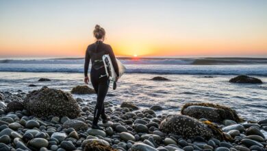 Woman wearing black neoprene surf booties walking across rocky beach toward ocean waves